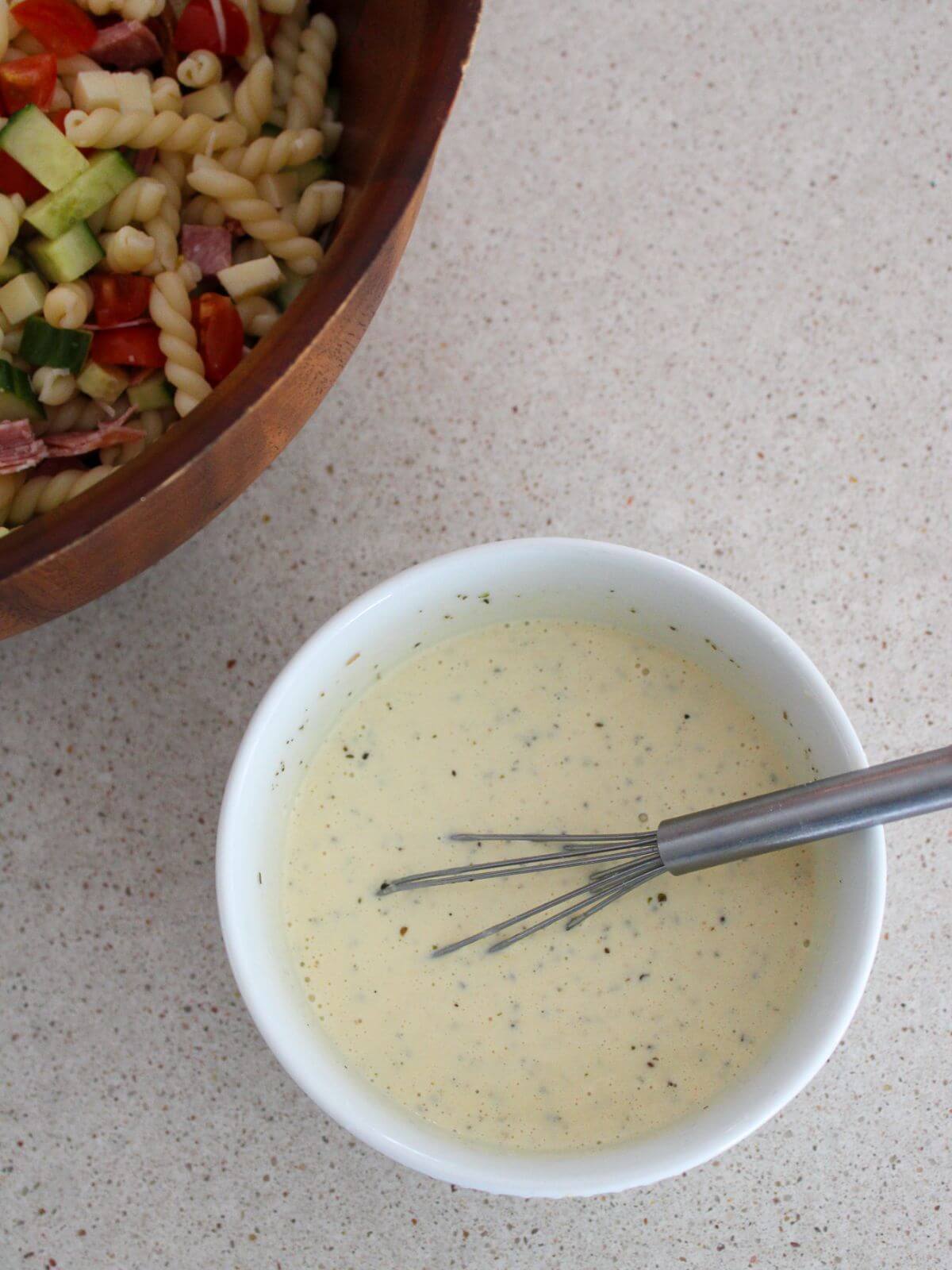 The creamy Italian dressing mixed in small white bowl with a silver spoon with the pasta salad next to it