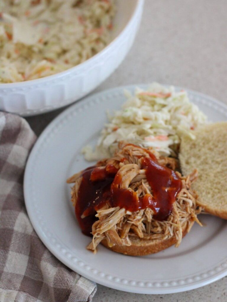 BBQ chicken on a bun on a plate with coleslaw and a plaid towel near