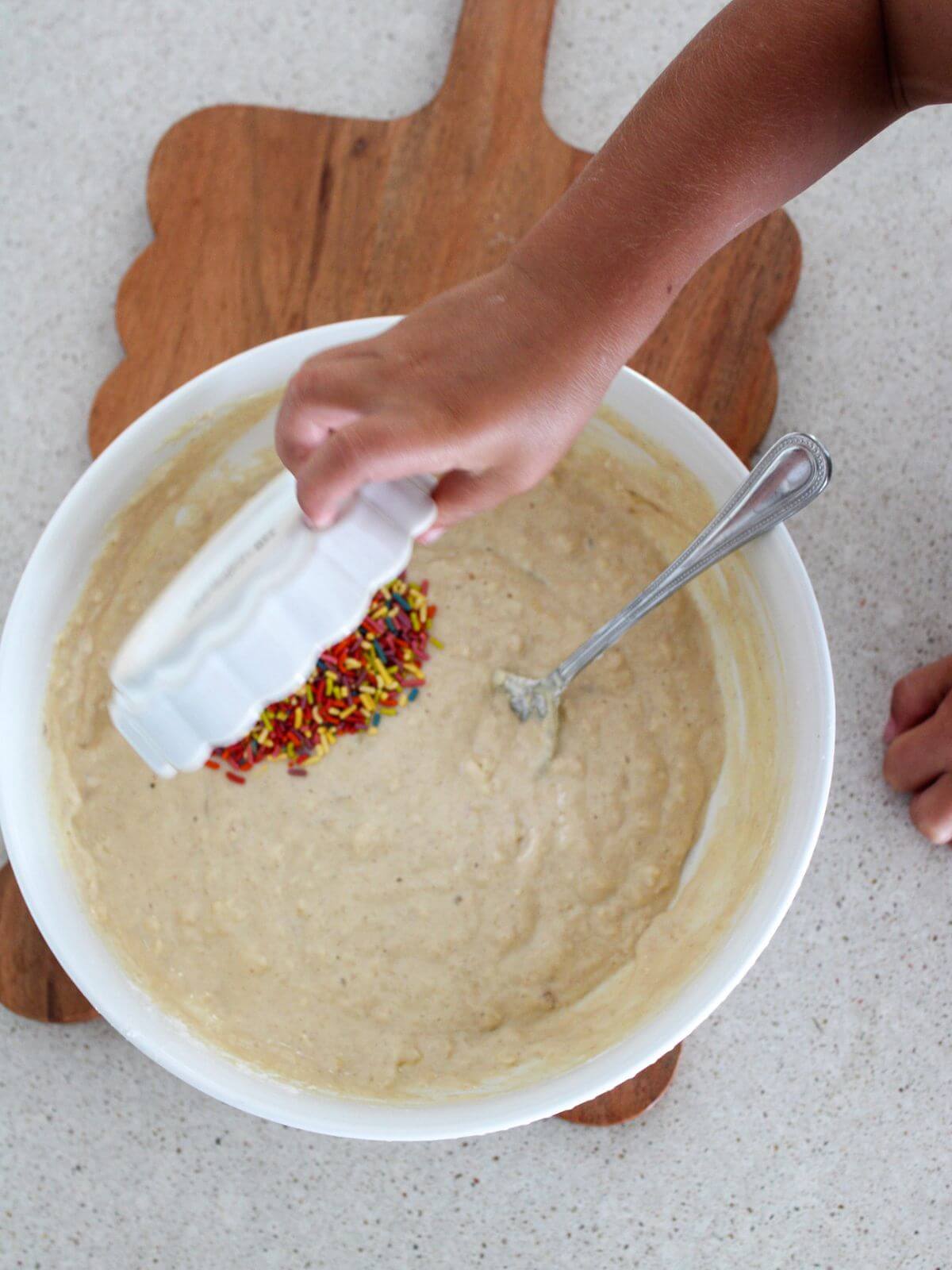 adding sprinkles to the batter in a white bowl with a silver spoon