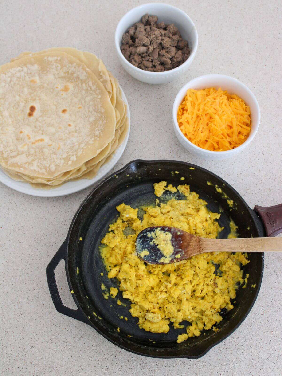 the ingredients ready to fill the burritos- eggs in a cast-iron pan, tortillas stacked on a white plate, cheese and sausage in white bowls