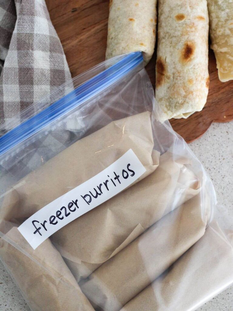 breakfast burritos wrapped in a freezer bag and a few scattered in the back on a cutting board with a plaid napkin