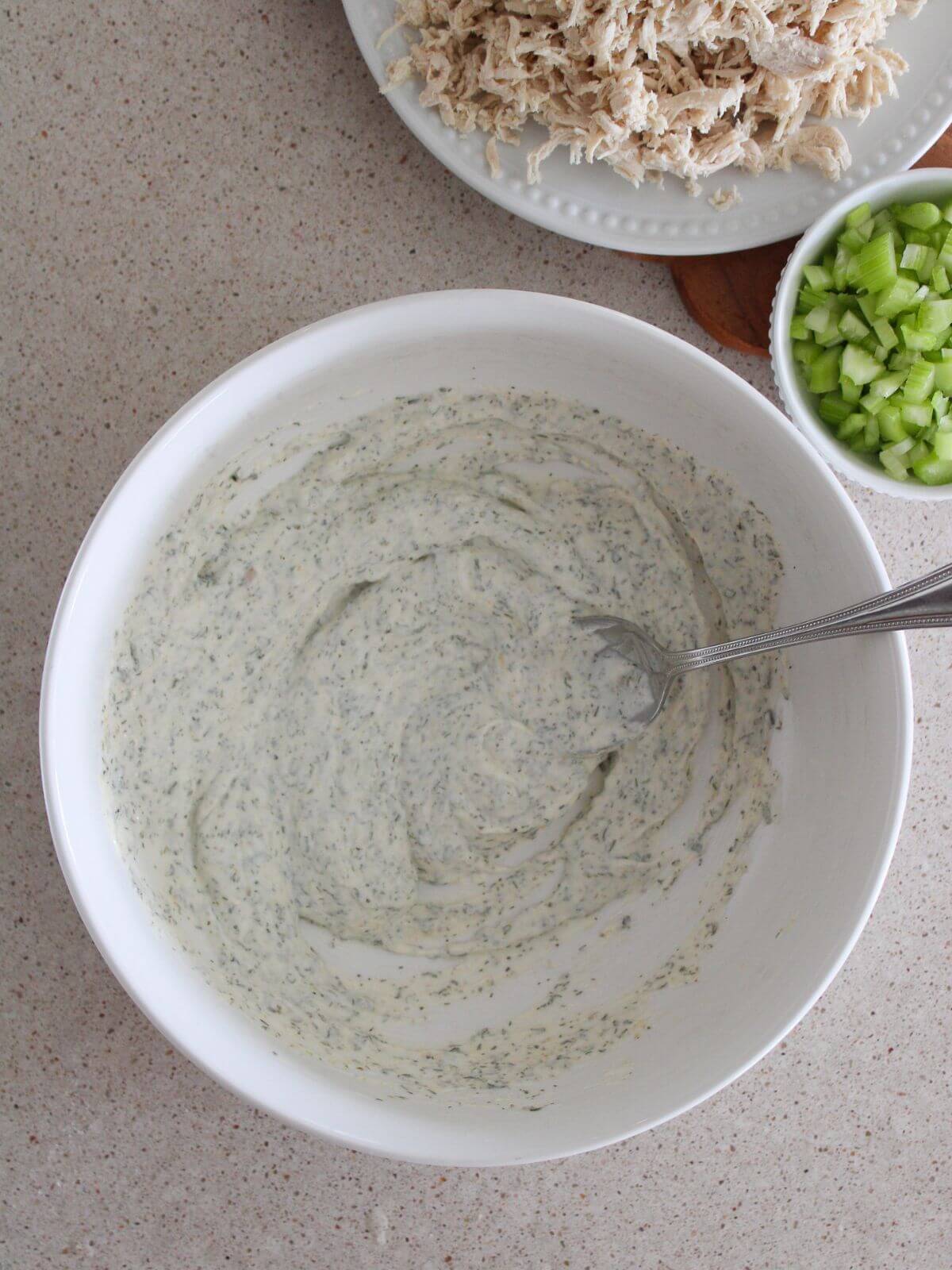 the dressing for the ranch chicken salad mixed together in a white bowl with a silver spoon with chicken and celery in the background