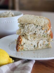 A high-protein ranch chicken salad sandwich stacked on a white plate with a lemon and plaid towel in front of it and a white bowl with extra chicken salad behind it