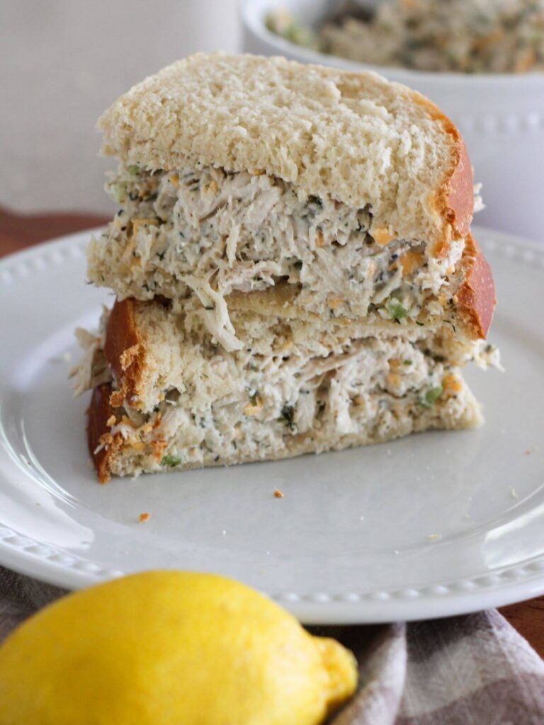 the high-protein ranch chicken salad on bread as a sandwich on a white plate with a lemon and plaid towel and a bowl of chicken salad behind it