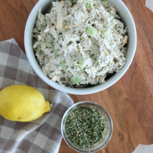 chicken salad in a white bowl with ranch seasoning, a lemon, and a plaid towel all on a cutting board