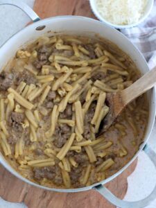 The One-Pot Creamy Ground Beef Pasta in the pot with a wooden spoon sitting on a cutting board with a small bowl of parmesan and a plaid napkin next to it