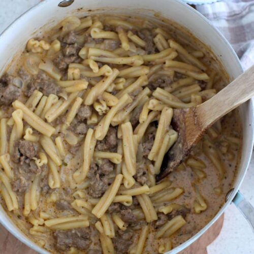 The One-Pot Creamy Ground Beef Pasta in the pot with a wooden spoon sitting on a cutting board with a small bowl of parmesan and a plaid napkin next to it