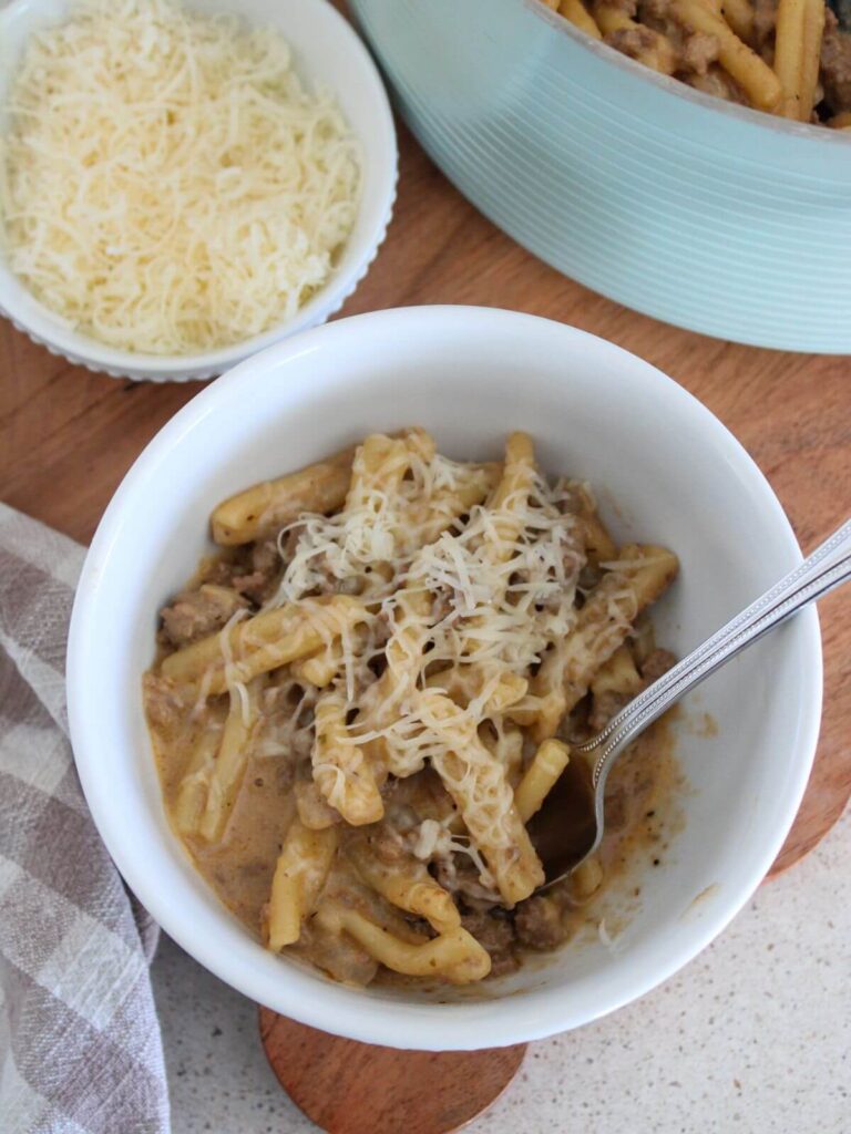 the pasta in a white bowl with a silver spoon on a cutting board with a plaid napkin, little bowl of parmesan and the rest of the pasta in the background