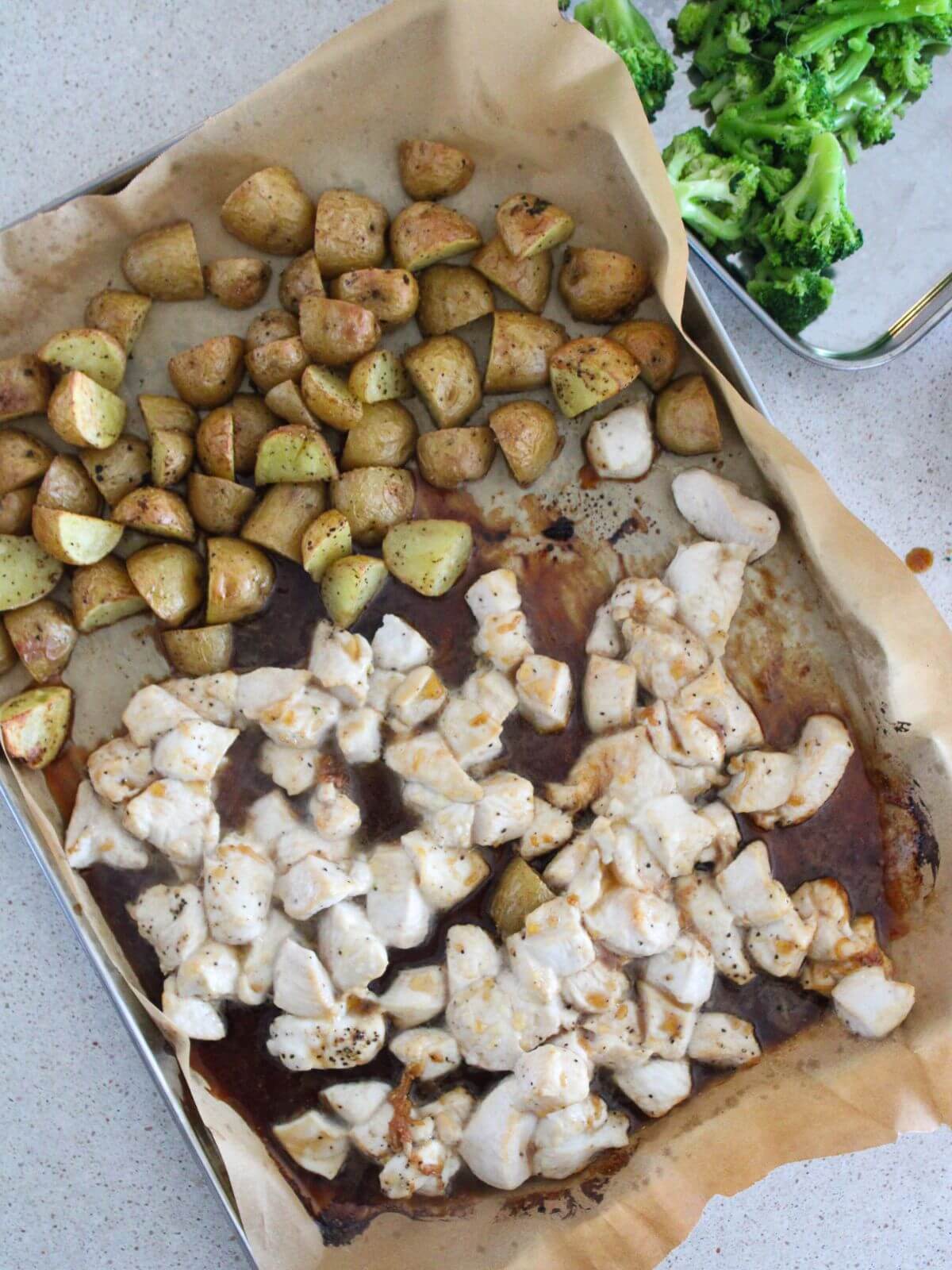 the chicken and potatoes roasted on a sheet pan with parchment paper the broccoli in the background