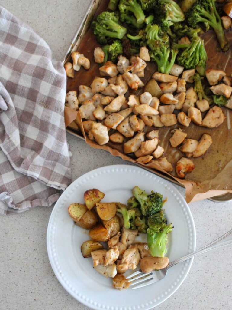 overhead view of the sheet pan chicken on the sheet pan in the background and a white plate plated with the meal in the front and a plaid towel next to it