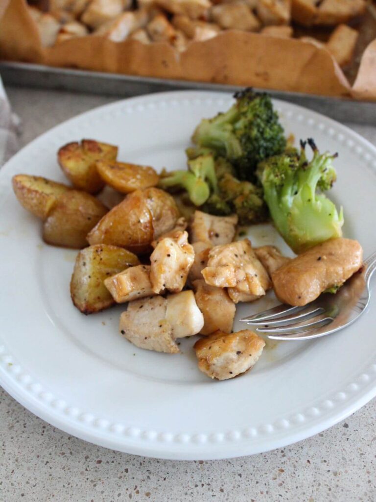 Up close of the honey garlic chicken plated on a white plate with a silver fork and the sheet pan with the rest of the meal in the background