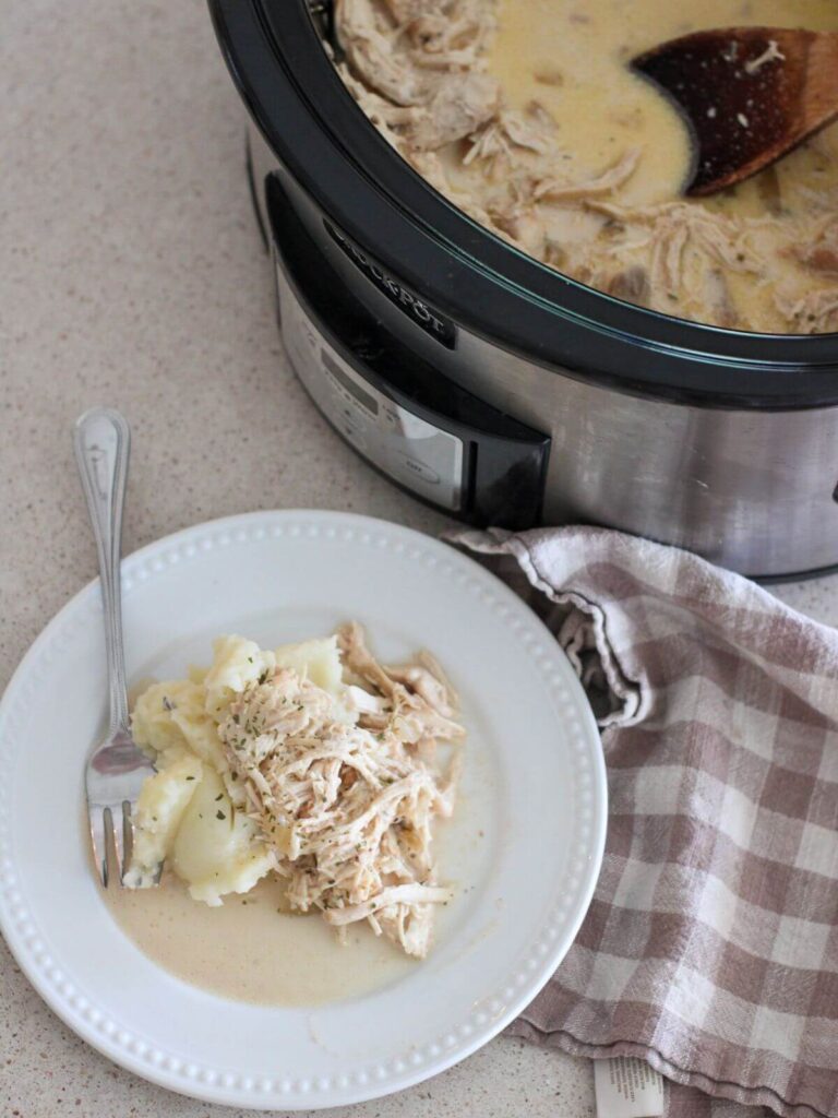 Easy Slow Cooker Chicken and Gravy on a white plate with a silver spoon with the rest in a crockpot behind with a plaid napkin