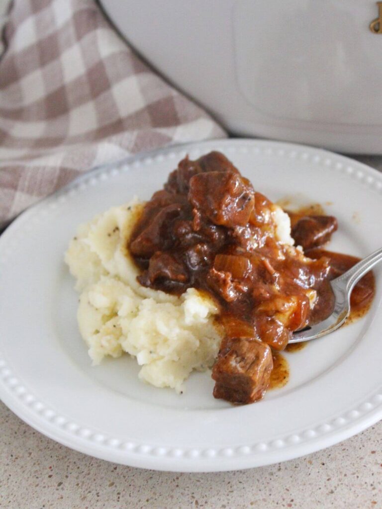 Slow Cooker Beef Tips and Gravy Plated on a white plate over mashed potatoes with a silver fork and plaid napkin the crockpot in the background