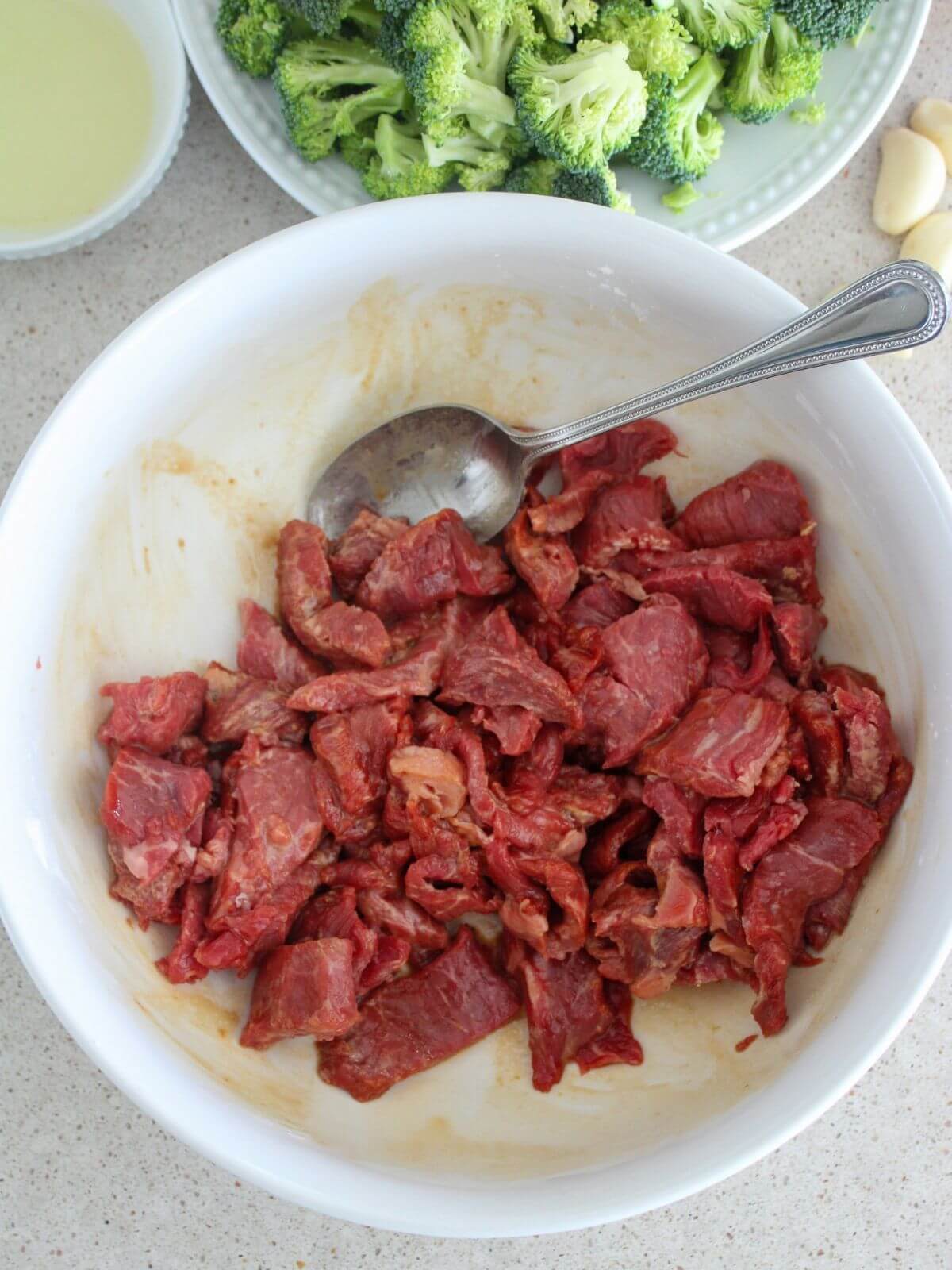 Beef marinating in a white bowl with a silver spoon with broccoli and olive oil behind