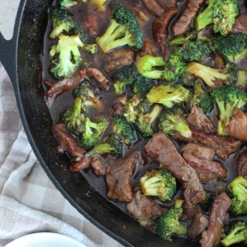 Easy Beef and Broccoli in a cast-iron pan on a cutting board with a plaid napkin and a small white bowl of green onions