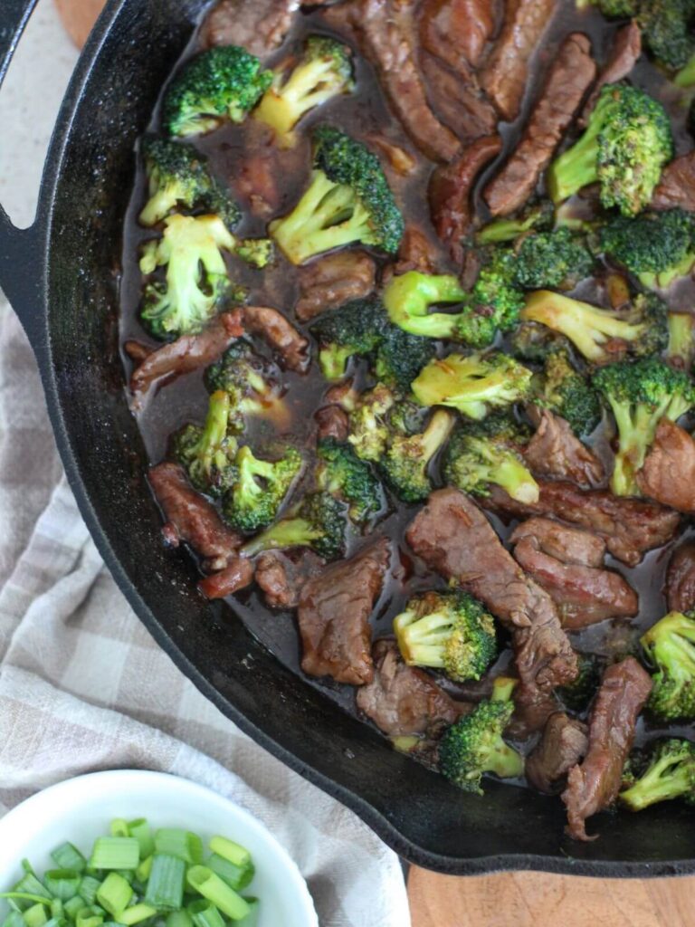 Easy Beef and Broccoli in a cast-iron pan on a cutting board with a plaid napkin and a small white bowl of green onions