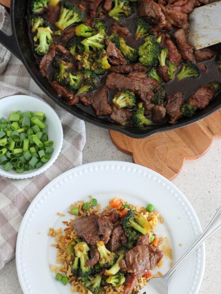 Beef and Broccoli plated on a white plate with fried rice and a silver spoon with the rest of the meal in a cast-iron pan behind with a plaid napkin and a small white bowl with green onions 