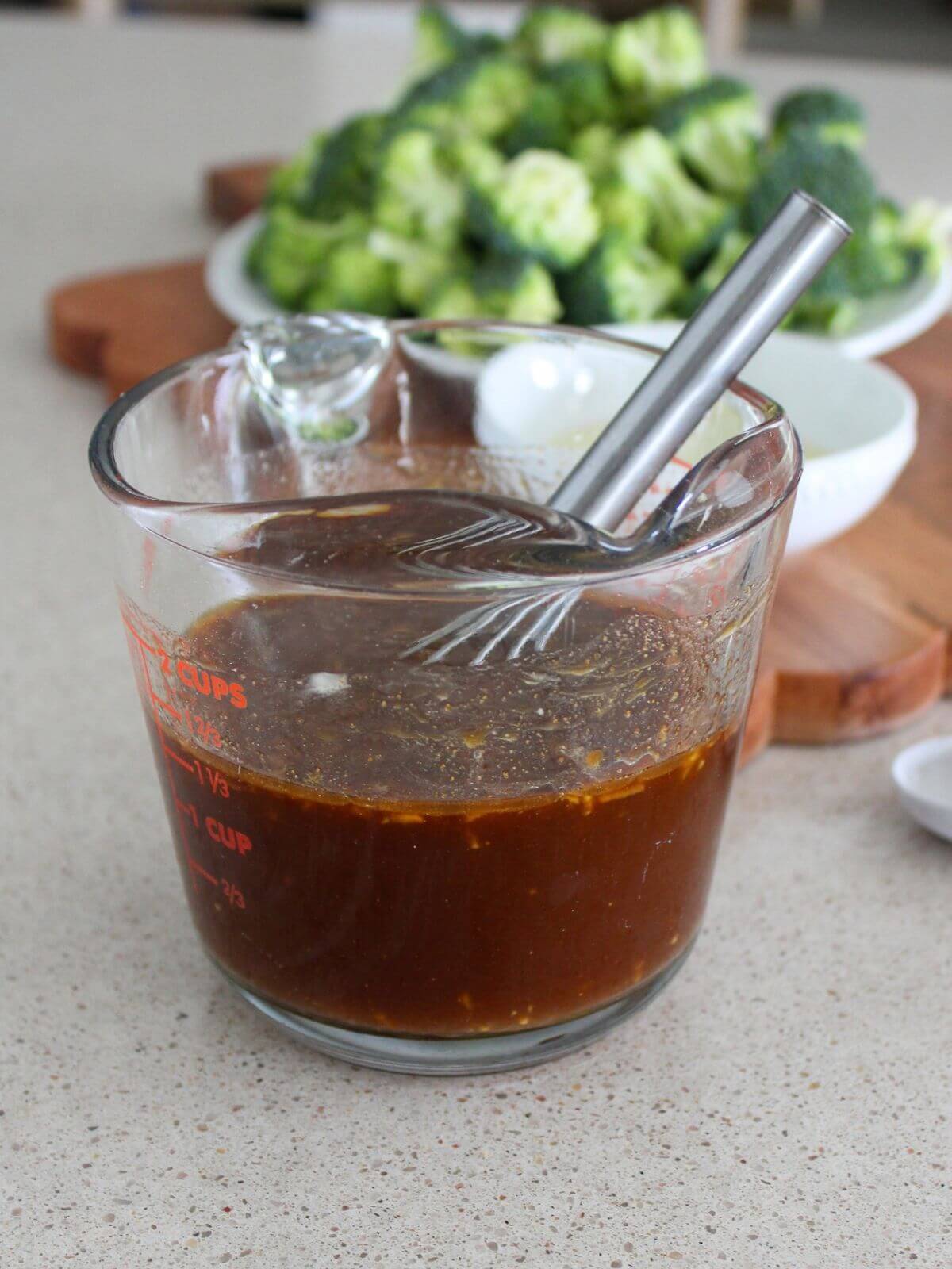 Mixing the sauce in a glass measuring cup with a silver whisk with cutting board and broccoli in the background