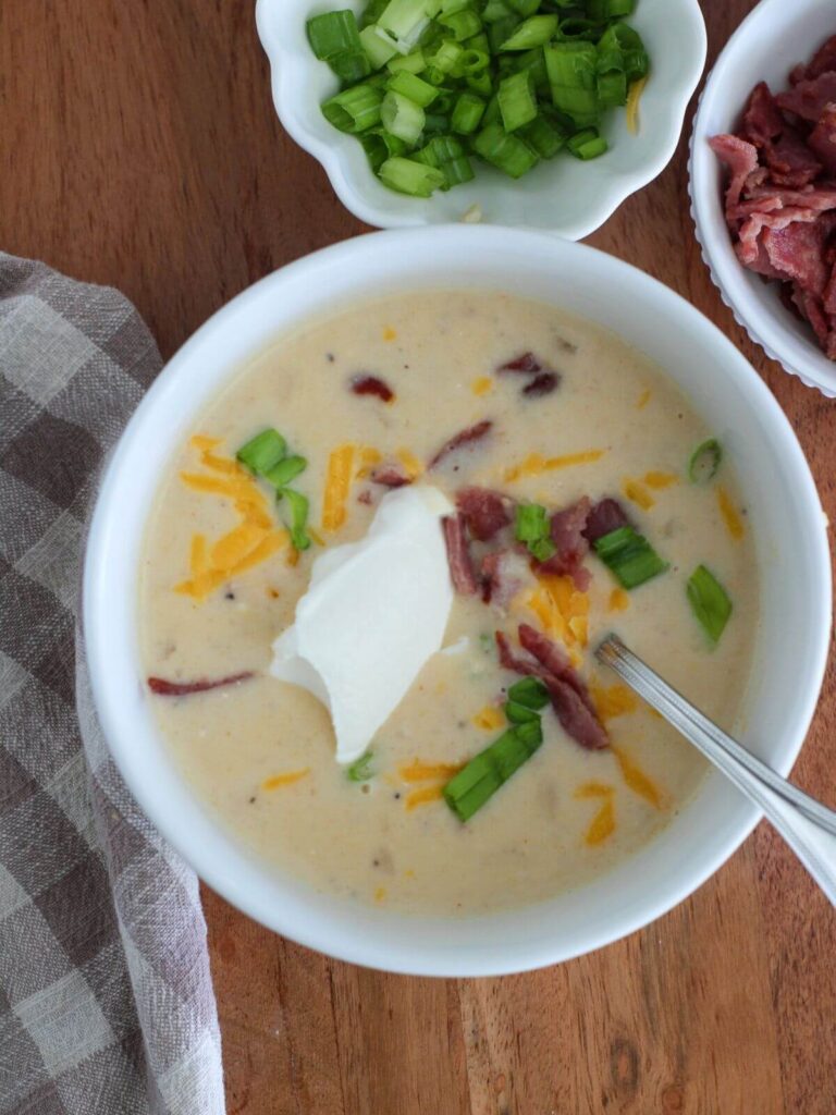 Loaded Potato Soup in bowl ready to eat on top of a cutting board with a plaid napkin and some small bowls with green onions and bacon close