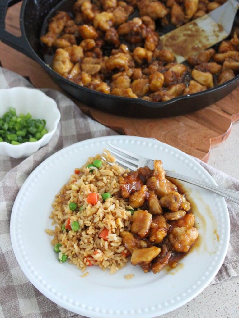 Better-Than-Takeout Orange Chicken on a white plate with fried rice and the cast-iron pan with the rest in the background on a wooden cutting board with a small white bowl of green onions and a plaid napkin