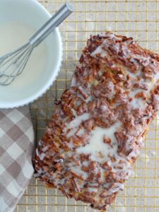 The apple fritter bread glazed on a golden cooling rack with a plaid napkin next to it and the glaze in a white bowl with silver whisk