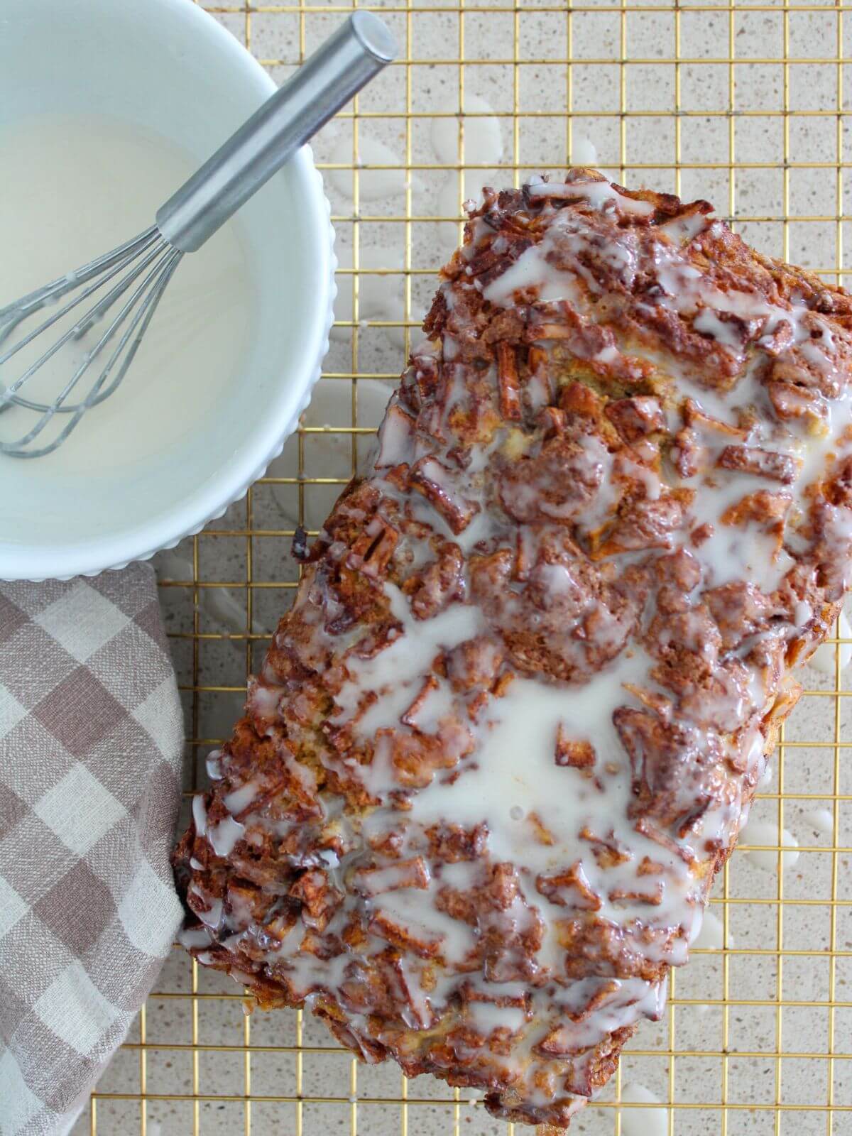 The apple fritter bread glazed on a golden cooling rack with a plaid napkin next to it and the glaze in a white bowl with silver whisk
