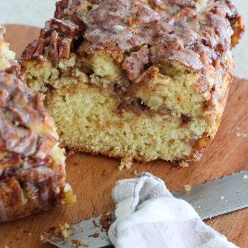 the apple fritter bread sliced on a wooden cutting board with a silver knife and a plaid napkin