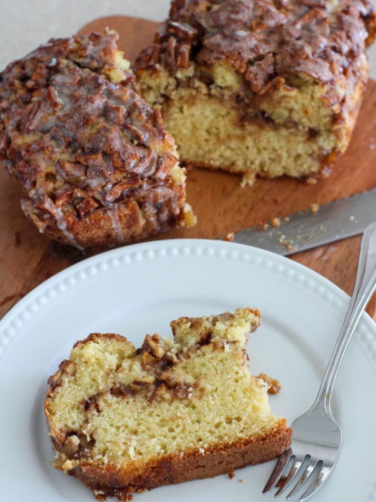 the apple fritter bread sliced on a cutting board with a silver bread knife and a piece on a white plate with a silver fork 