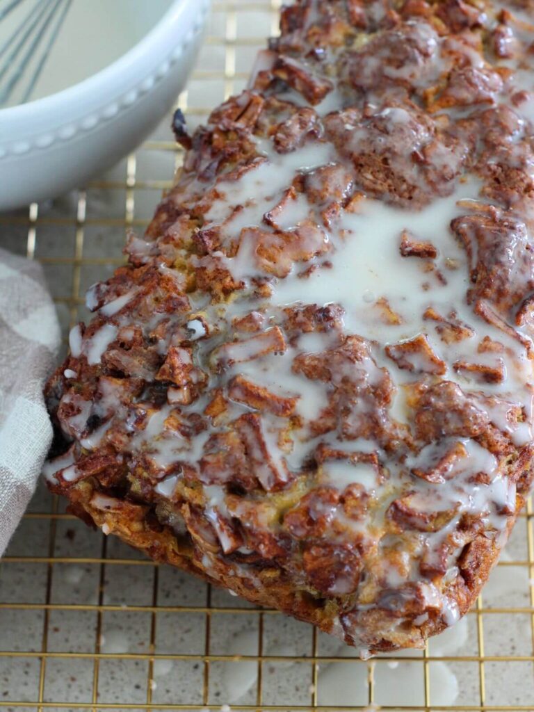 Easy Apple Fritter Bread glazed on a cooling rack with a plaid towel and the rest of the glaze in a white bowl with a whisk next to it