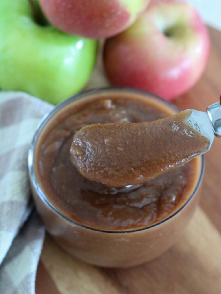 the apple butter in a glass jar with the apple butter on a serving knife on a cutting board with apples and a plaid napkin around it