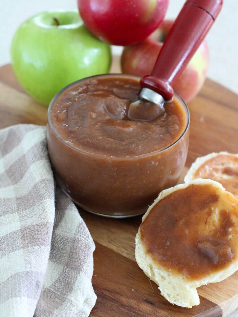 Homemade Apple Butter in a glass jar with a serving spoon on a cutting board with apples and a biscuit with apple butter on it with a plaid napkin