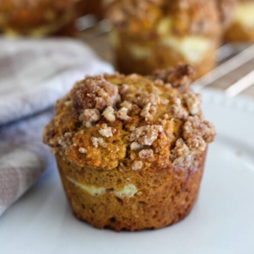 A close up on one pumpkin muffin on a white plate with a plaid napkin and the rest of the muffins behind on a cooling rack