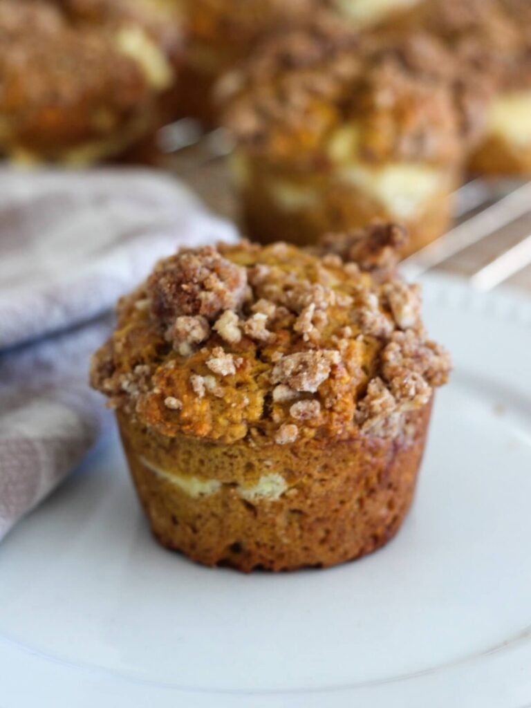 A close up on one pumpkin muffin on a white plate with a plaid napkin and the rest of the muffins behind on a cooling rack