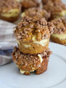 Pumpkin Cream Cheese Muffins stacked on top of each other on a white plate with a plaid napkin and the rest of the muffins behind