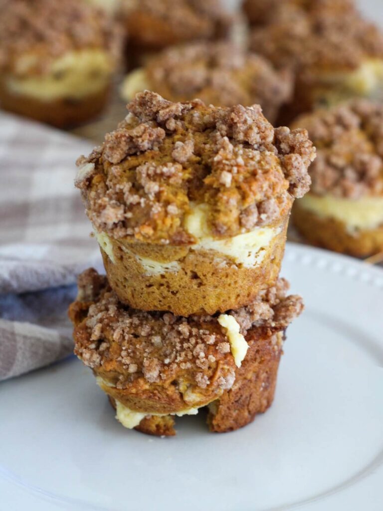 Pumpkin Cream Cheese Muffins stacked on top of each other on a white plate with a plaid napkin and the rest of the muffins behind